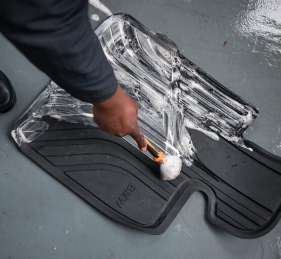 A person uses a brush to clean a car floor mat, emphasising the significance of thorough vehicle maintenance and detailing services.